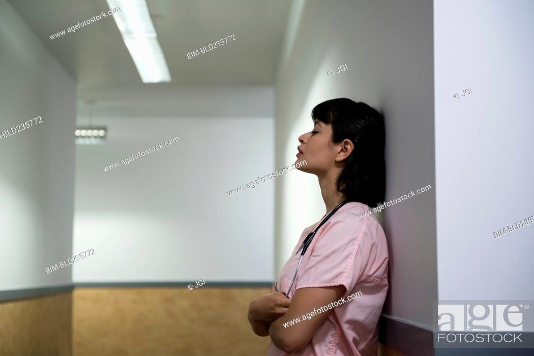 Stock Photo: Tired Hispanic nurse leaning on wall in hospital corridor.