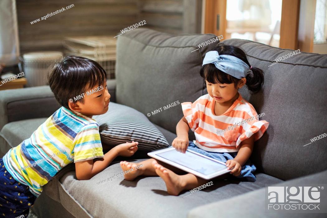 Stock Photo: Young girl with black pigtails sitting on a sofa, digital tablet on her lap, boy standing beside her.