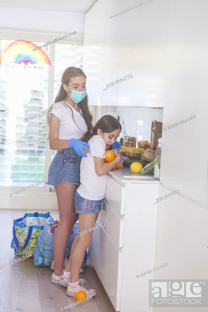 Photo de stock: little girl helps sister (that is wearing mask and gloves) in the kitchen.