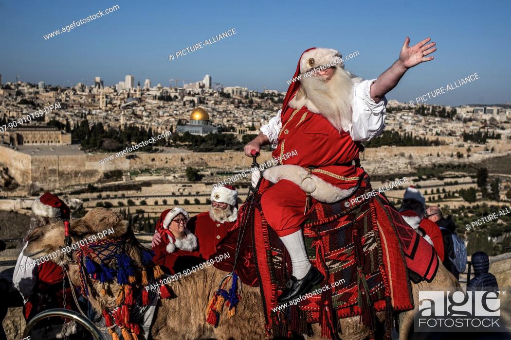 Stock Photo: 07 January 2020, ---, Jerusalem: A man dressed up as Santa Claus wave as he rides a camel on the Mount of Olives backdropped by Jerusalem's Old City and the.