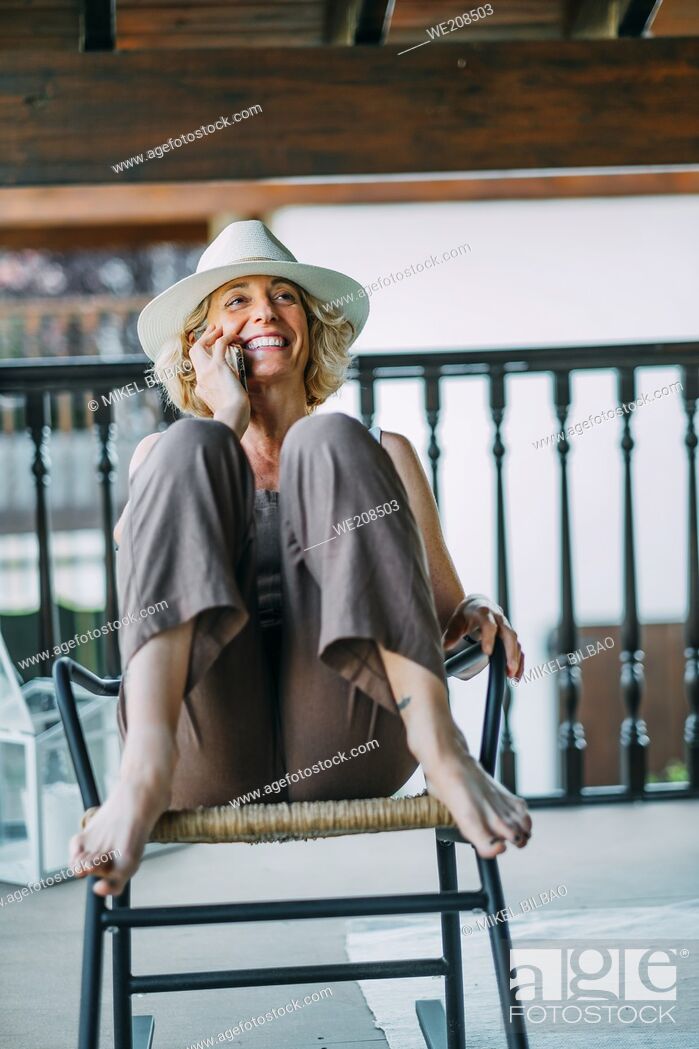 Stock Photo: Portrait of a mature caucasian blonde young woman in her 50s talking on a mobile phone smiling and wearing a hat and sitting in a relaxed pose in a rocking.