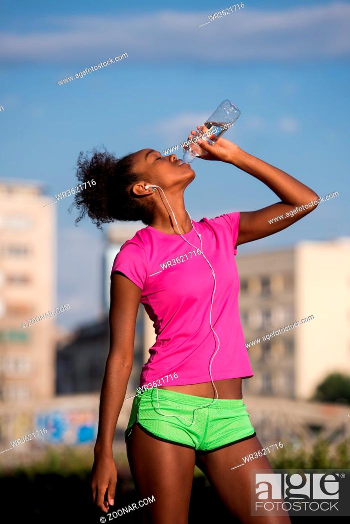 Photo de stock: Young beautiful african american woman with headphones drinking water after jogging in the city.