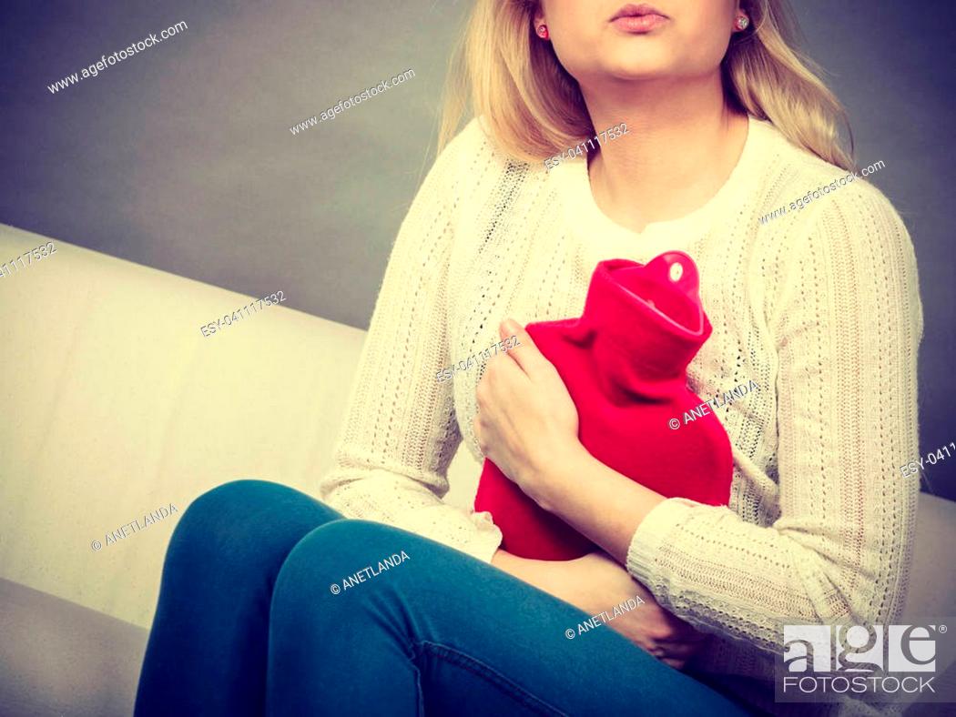 Stock Photo: Painful periods and menstrual cramp problems concept. Woman having stomach cramps sitting on cofa feeling very unwell holding hot water bottle to feel some.