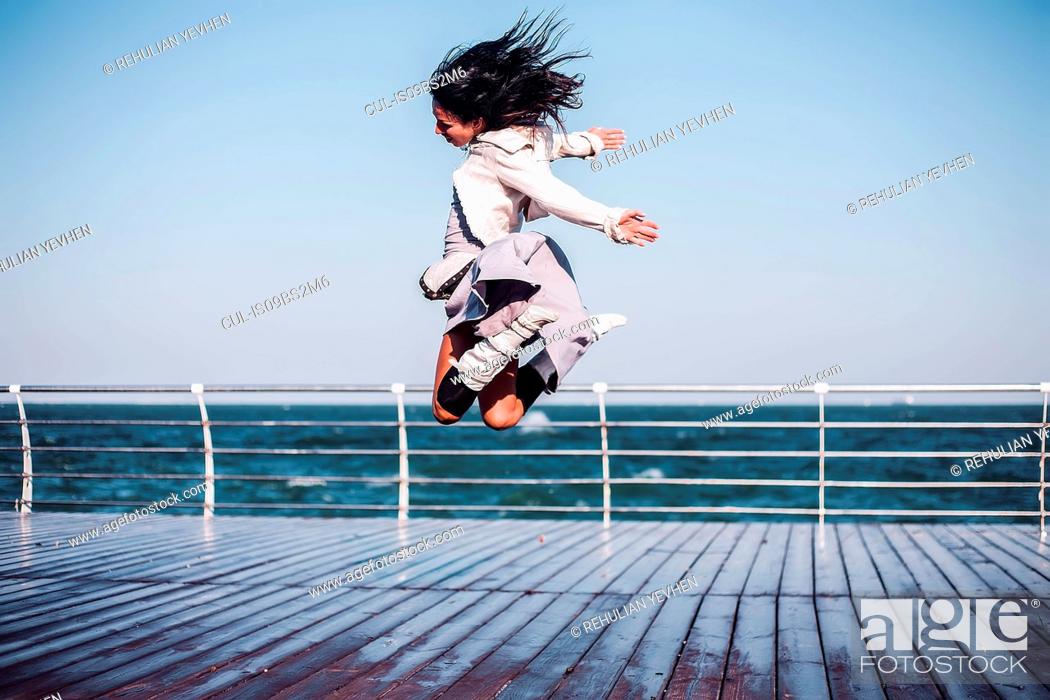 Stock Photo: Mid adult woman jumping mid air on sea pier, Odessa, Odeska Oblast, Ukraine.