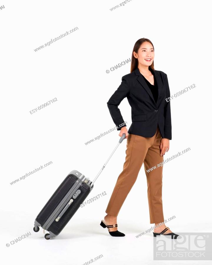 Imagen: Young asian women in black suit smiles as she walks dragging a black suitcase. Portrait on white background with studio light.