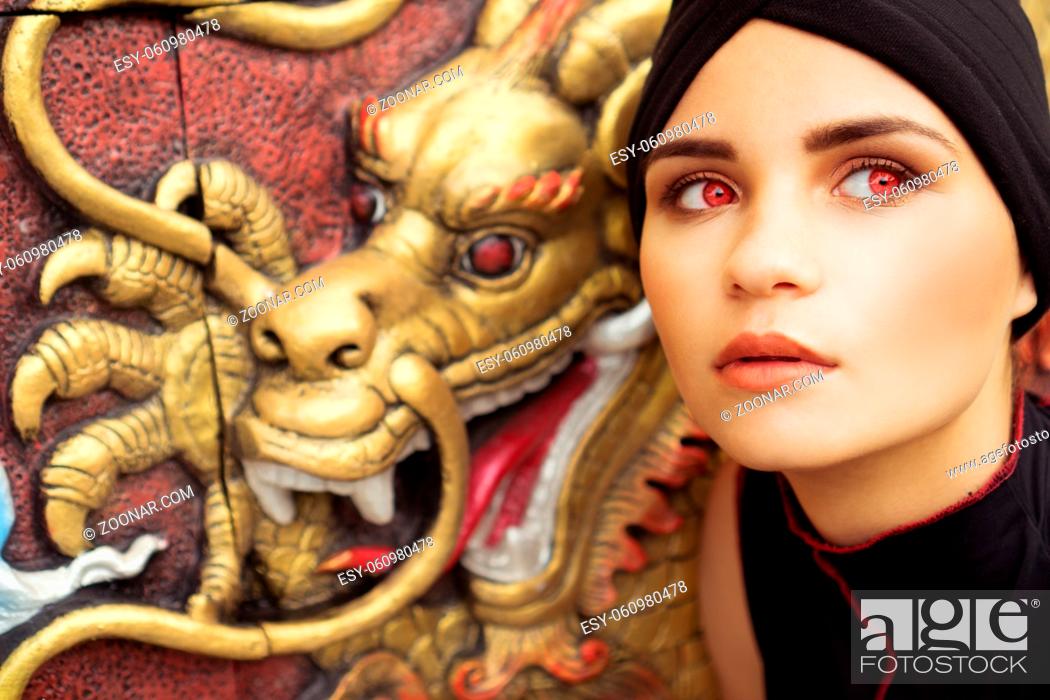 Stock Photo: Fashion closeup portrait of young brunette woman with black hat and red eyes over golden dragon on the wall background.