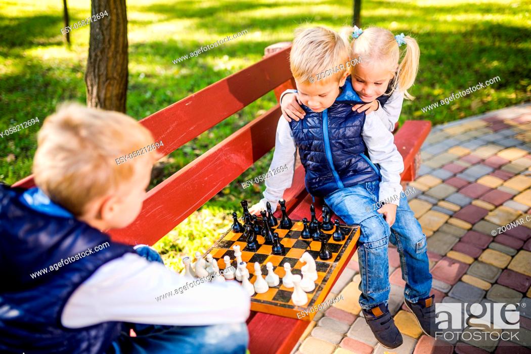 Stock Photo: Caucasian children play chess on wooden chessboard in park bench. Brothers and sister big friendly active mental family spend time on vacation in sunny weather.