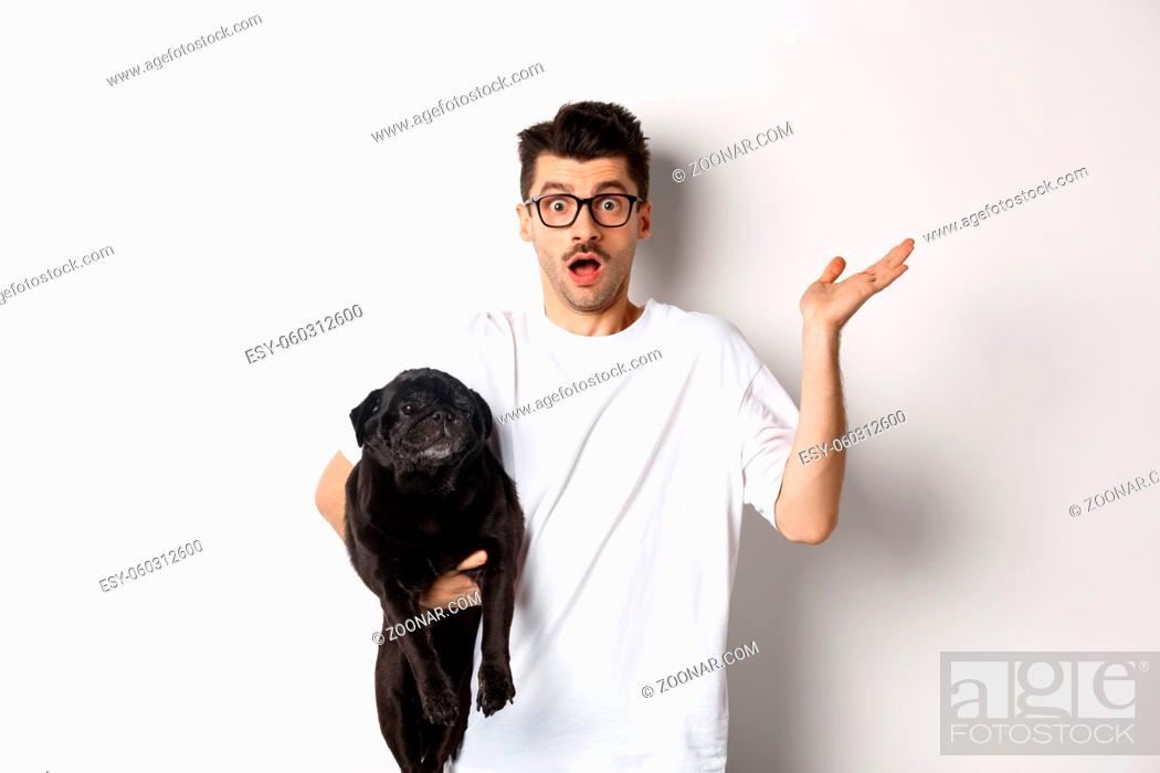 Stock Photo: Image of confused hipster man holding dog and shrugging, dont know, raising hand puzzled, standing with his animal over white background.
