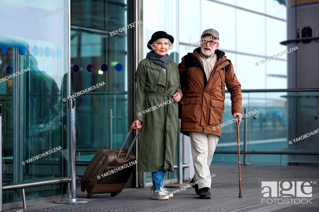 Stock Photo: Arrival. Full length portrait of pleasant positive senior man with cane and woman with suitcase are standing together near airport building.