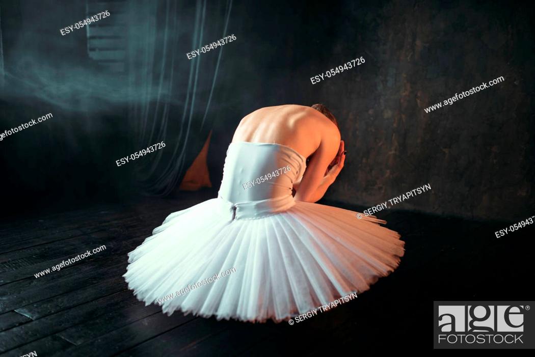 Imagen: Classical ballet dancer in white dress sitting on theatrical stage, back view. Ballerina training in class with black background.