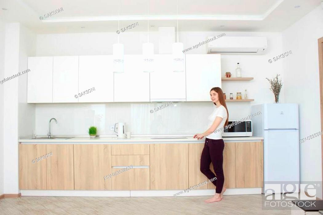 Stock Photo: Pretty young woman In a white T shirt standing in kitchen and smiling. Real interior. Wide angle.