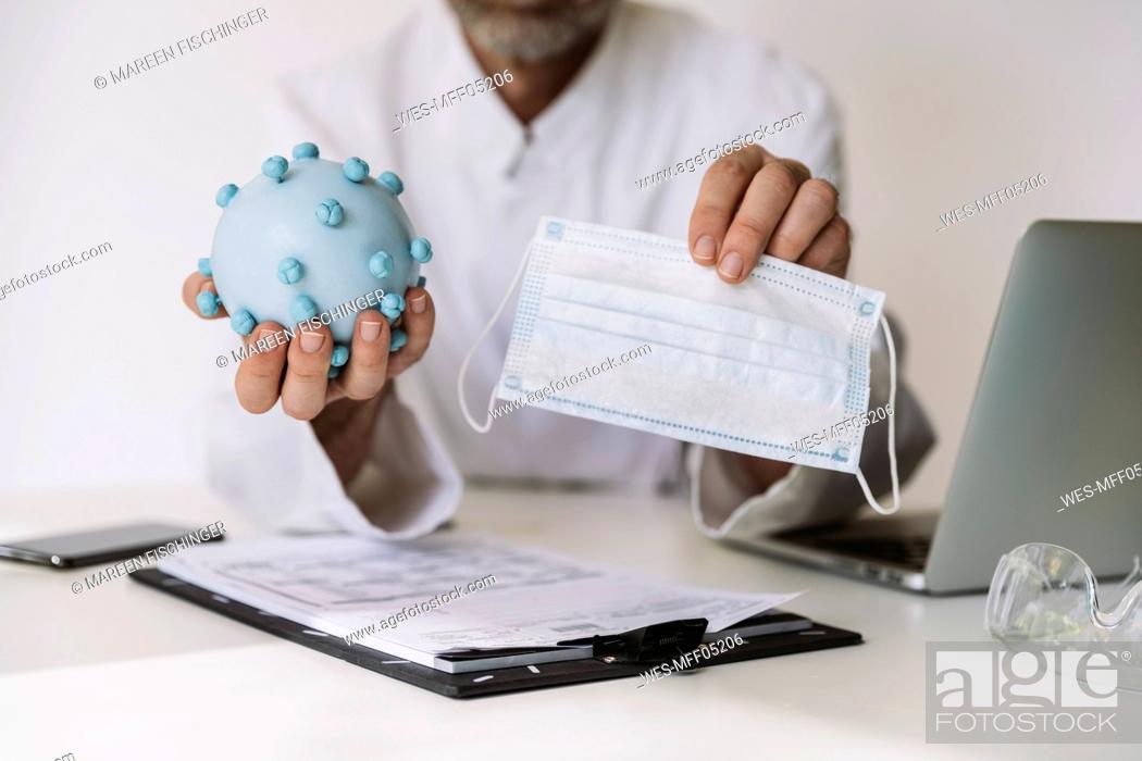 Imagen: Close-up of scientist holding corona virus model and mask at desk.