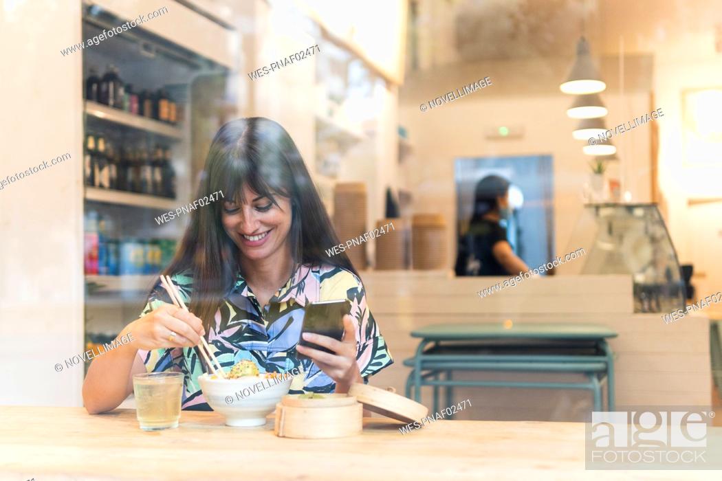 Stock Photo: Smiling woman with smart phone having food at restaurant seen through glass.
