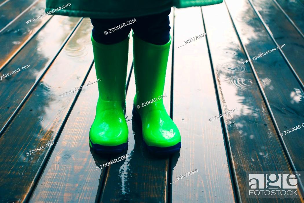 Stock Photo: Closeup of legs of kid standing on a porch wearing green wellies and raincoat. Rain boots in bright green color.