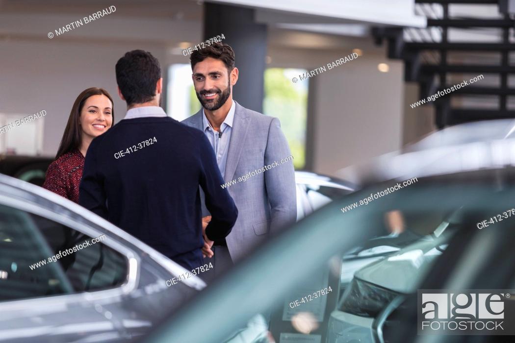 Stock Photo: Car salesman greeting, shaking hands with couple customers in car dealership showroom.