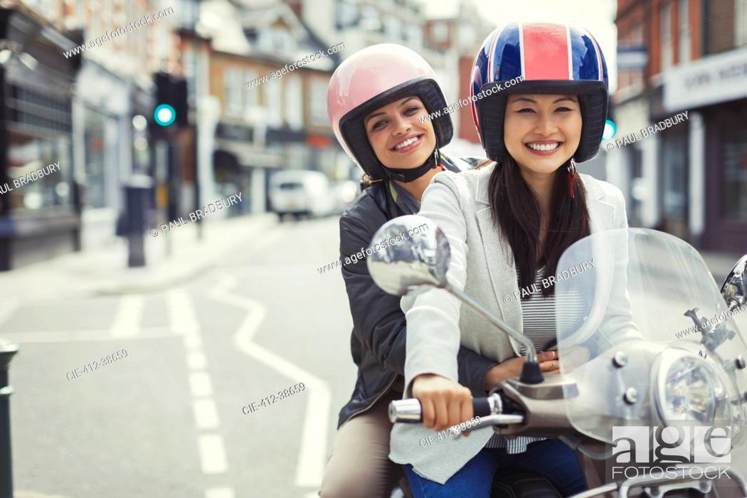 Imagen: Smiling young women friends wearing helmets, riding motor scooter on urban street.