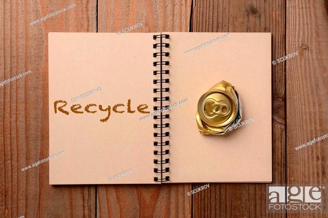 Stock Photo: A crushed soda can on the blank page of a notebook. The opposite page has the word Recycle spelled out. Horizontal format on a rustic wooden table.