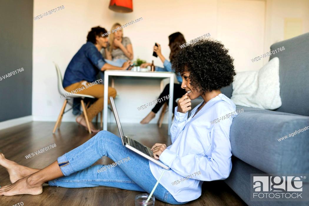 Stock Photo: Woman sitting on floor using laptop with friends in background.