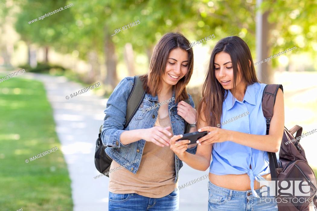 Stock Photo: Two beautiful young ethnic twin sisters with backpacks using A smartphone outside.