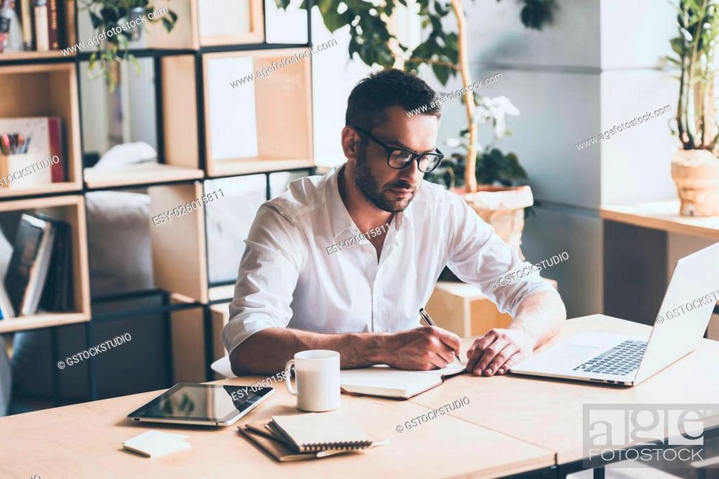 Stock Photo: Making some notes. Concentrated mature man writing something in note pad while sitting at his working place in office.