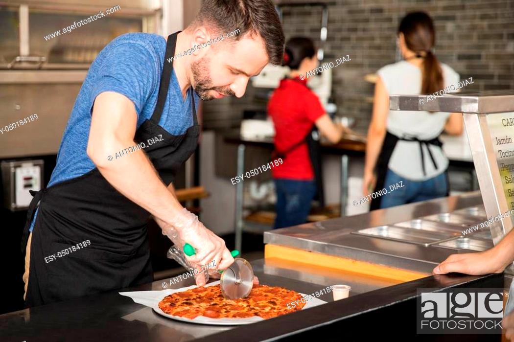 Imagen: Handsome man wearing plastic hand gloves and black apron using roller cutter to cut pizza in slices at pizza shop counter.