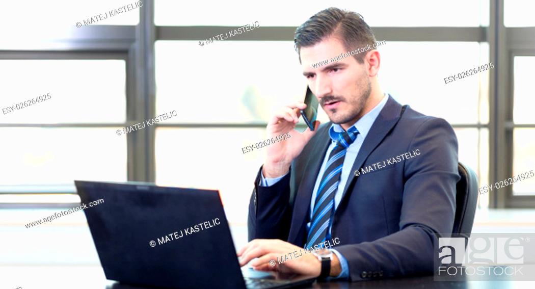 Imagen: Portrait of successful corporate businessman in bright modern office focused on data on his laptop computer while talking on mobile phone.