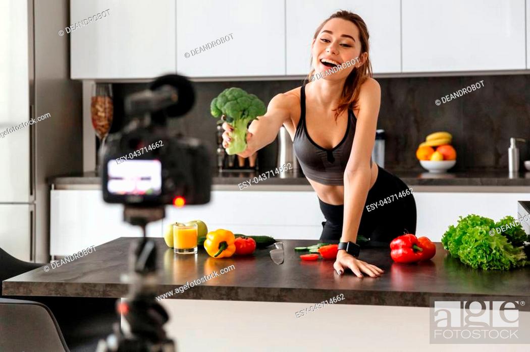 Stock Photo: Cheerful healthy young girl recording her blog episode about healthy food diet while standing at the kitchen at home.