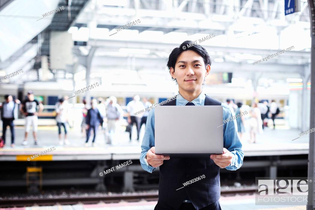 Stock Photo: Businessman wearing blue shirt and vest standing on train station platform, holding laptop, looking at camera.