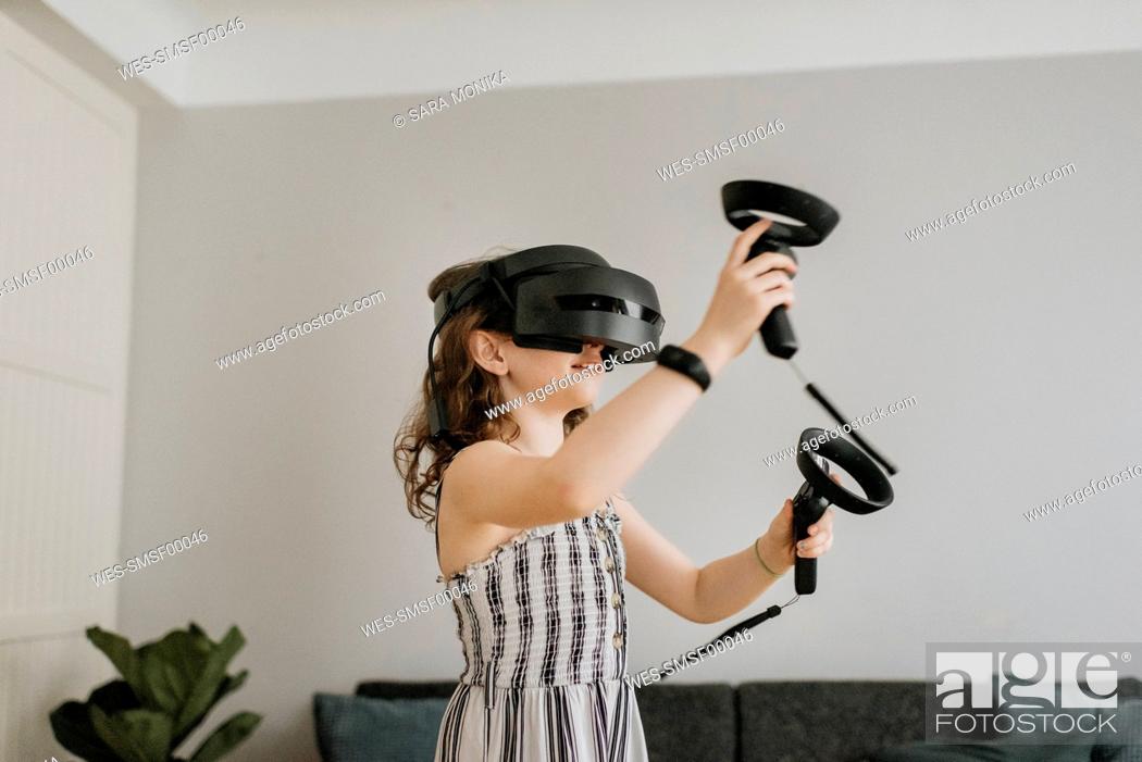 Photo de stock: Girl wearing virtual reality simulator playing with joysticks at home.