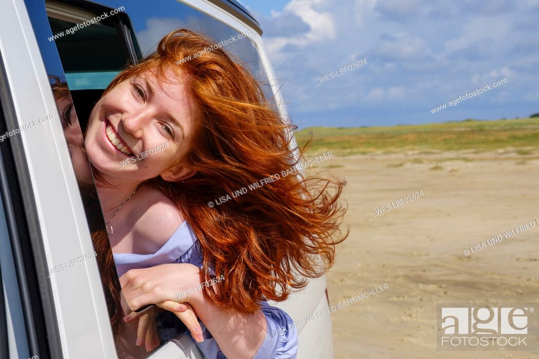 Stock Photo: Portrait of carefree redhead teenage girl with head out of car window at beach against sky.