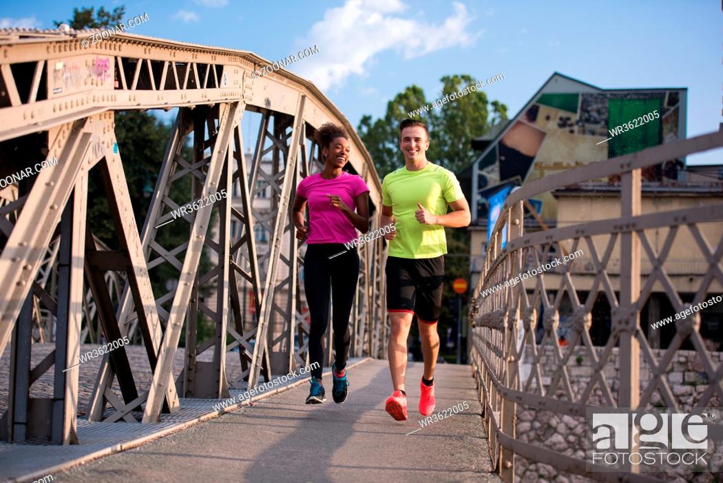 Photo de stock: healthy young multiethnic couple jogging across the bridge in the city at early morning with sunrise in background.