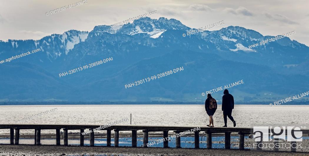 Stock Photo: 13 January 2023, Bavaria, Seebruck: Two men walk over a footbridge in Lake Chiemsee in spring-like temperatures against the almost snow-free backdrop of the.