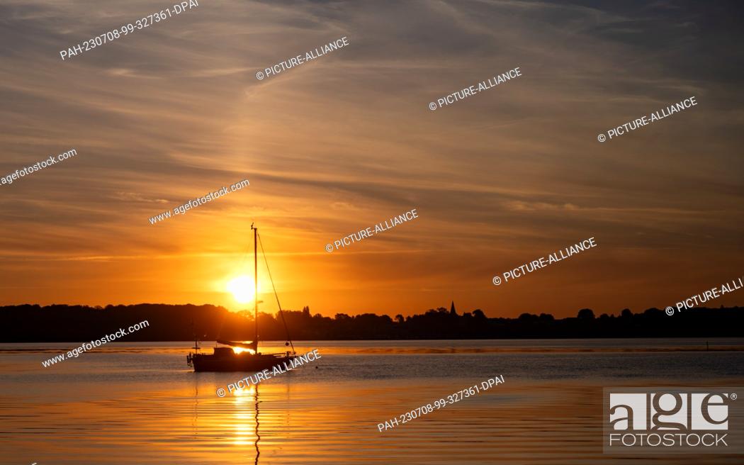 Stock Photo: 08 July 2023, Mecklenburg-Western Pomerania, Stralsund: The sun rises over the island of Rügen and bathes the sky over the Stralsund off the island of Rügen in.