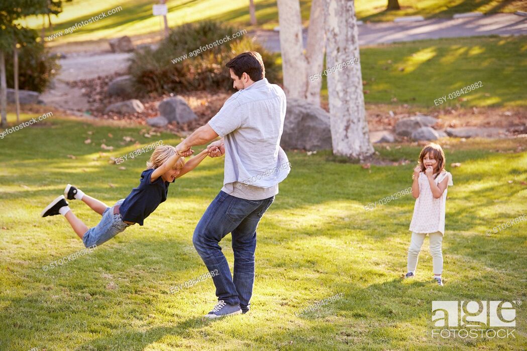Stock Photo: Father Swinging Son By His Arms In Park As Daughter Watches.