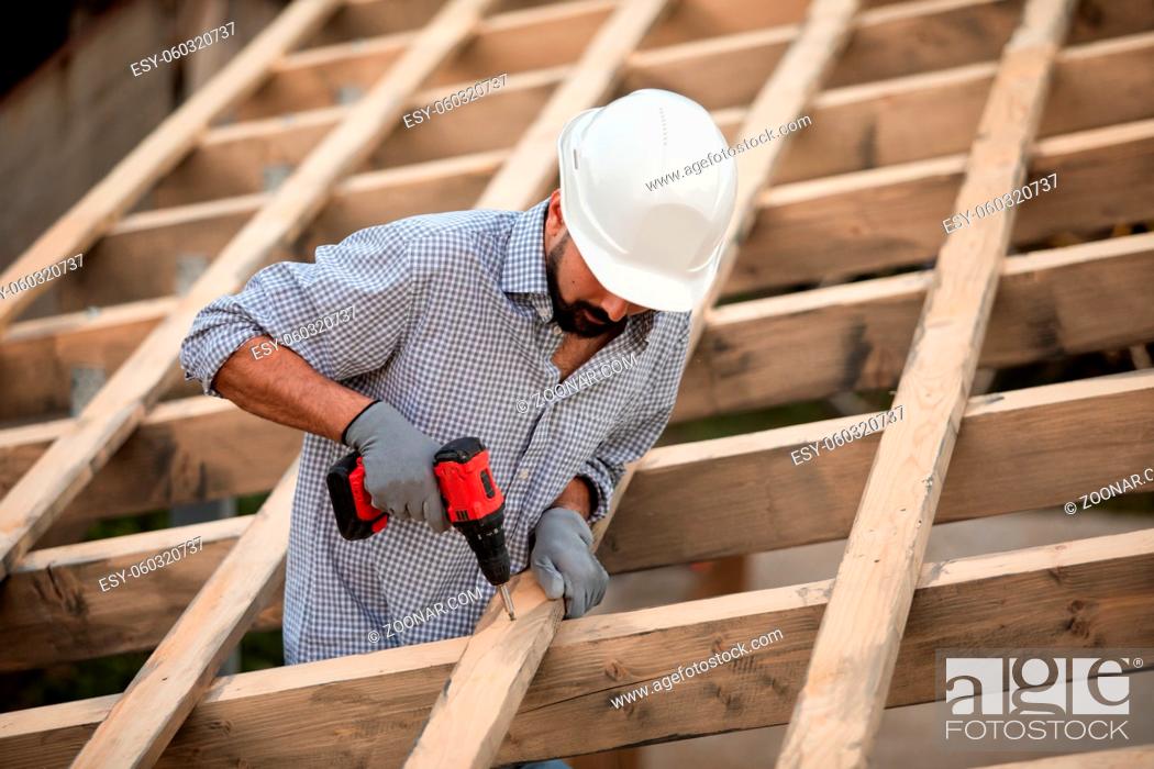 Stock Photo: The male worker in a protective helmet and gloves works on the roof using a screwdriver. He finishes the wooden frame on the roof.