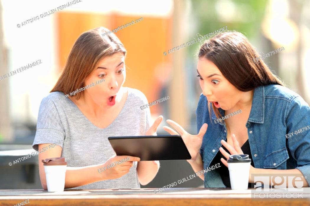 Stock Photo: Two amazed friends watching media content in a tablet sitting in a park.