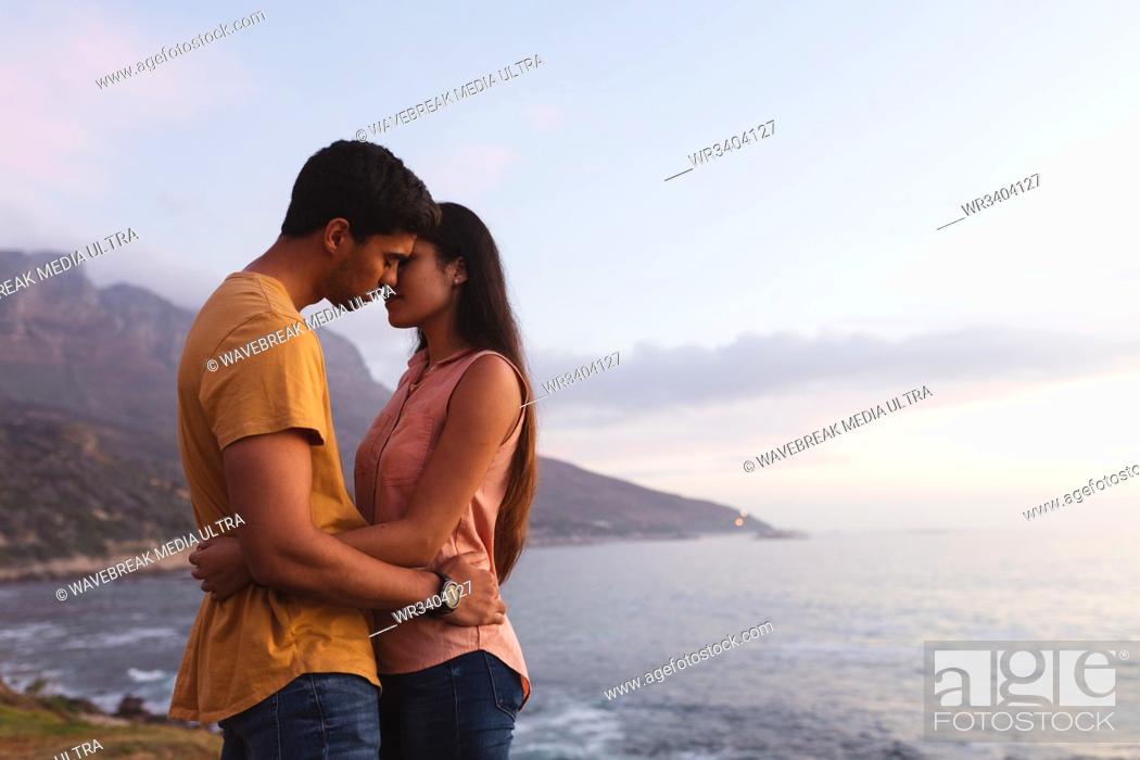 Stock Photo: Side view of a young mixed race couple standing on a beach embracing and about to kiss at sundown.