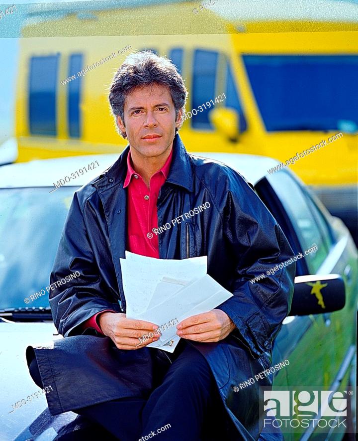 Stock Photo: Italian song writer Claudio Baglioni (Claudio Enrico Paolo Baglioni) sittimg on a car's hood holding some letters. On the background there is the yellow van.
