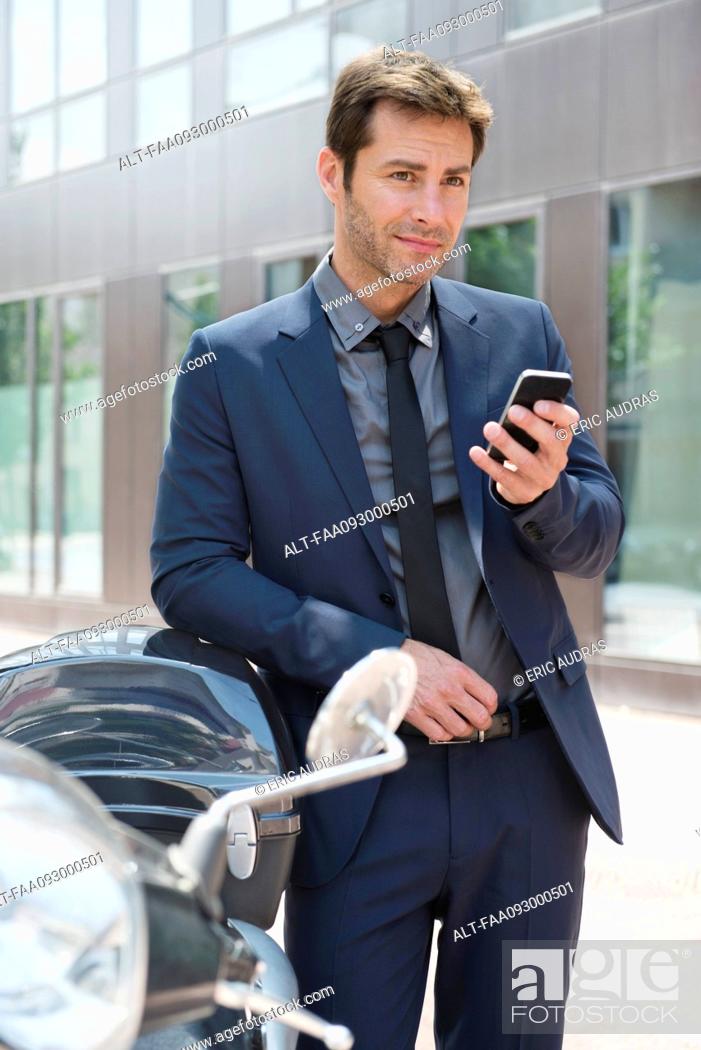 Stock Photo: Businessman using smartphone in city, leaning against motor scooters.