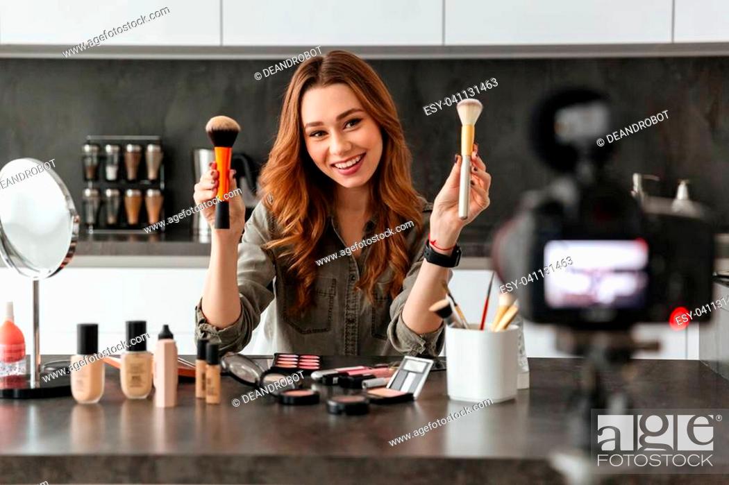 Stock Photo: Cheerful young girl recording her video blog episode about new cosmetic products while sitting at the kitchen table at home and applying make-up.