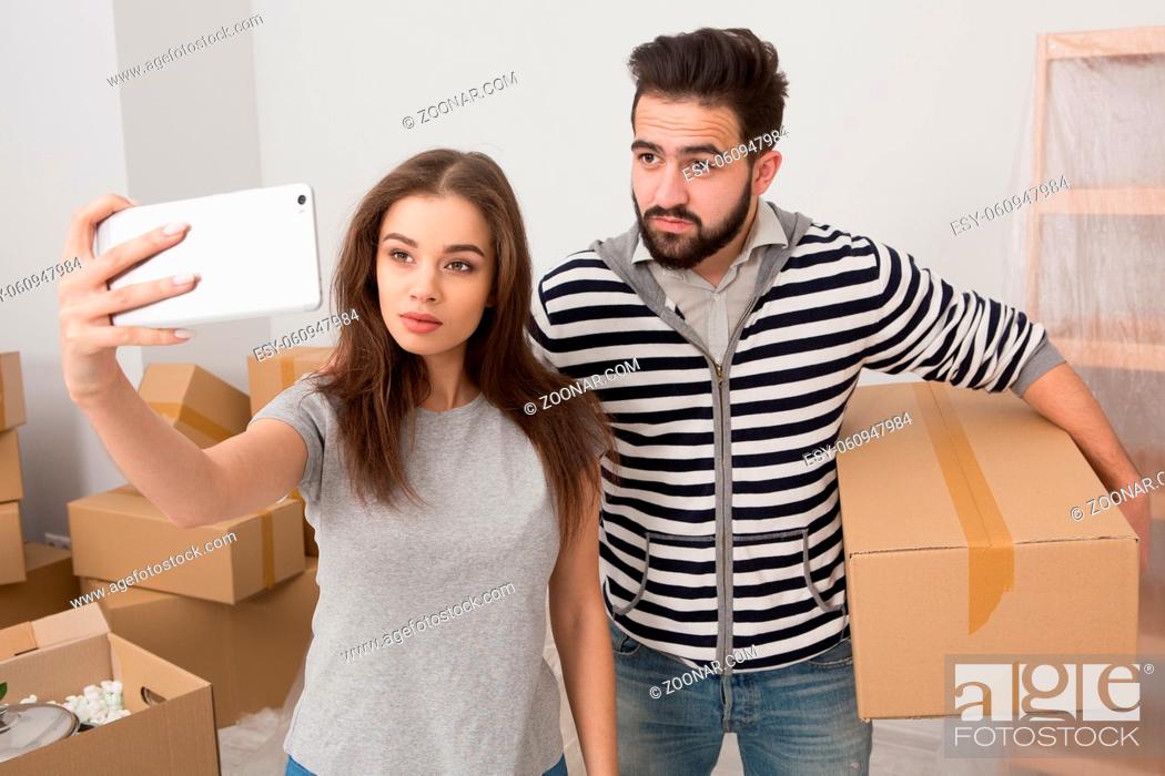 Stock Photo: Relocation young positive couple making selfie shot in new home. Man and woman in the room full of unpacked boxes, man holding box and woman holding phone.
