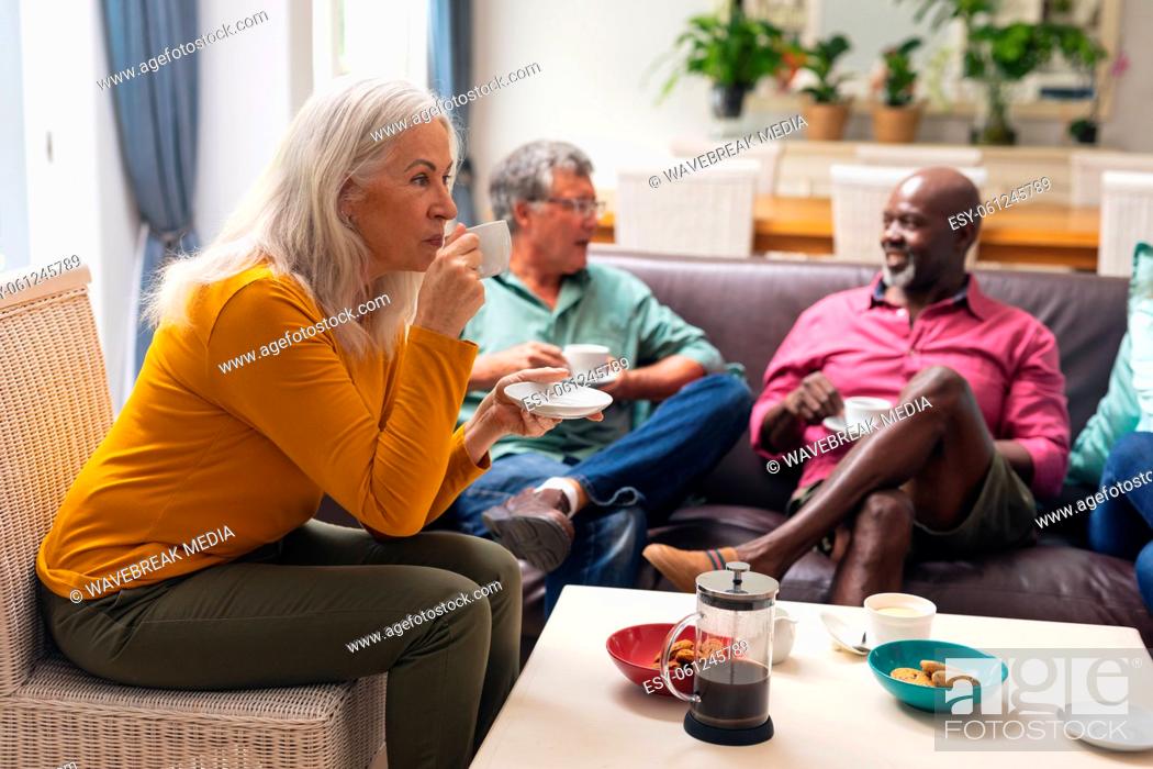 Stock Photo: Senior woman drinking coffee while multiracial friends talking in background at home.