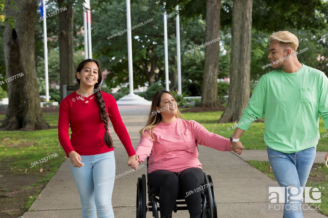 Stock Photo: Young woman in a wheelchair holds hands with the friends that are walking down a path beside her.