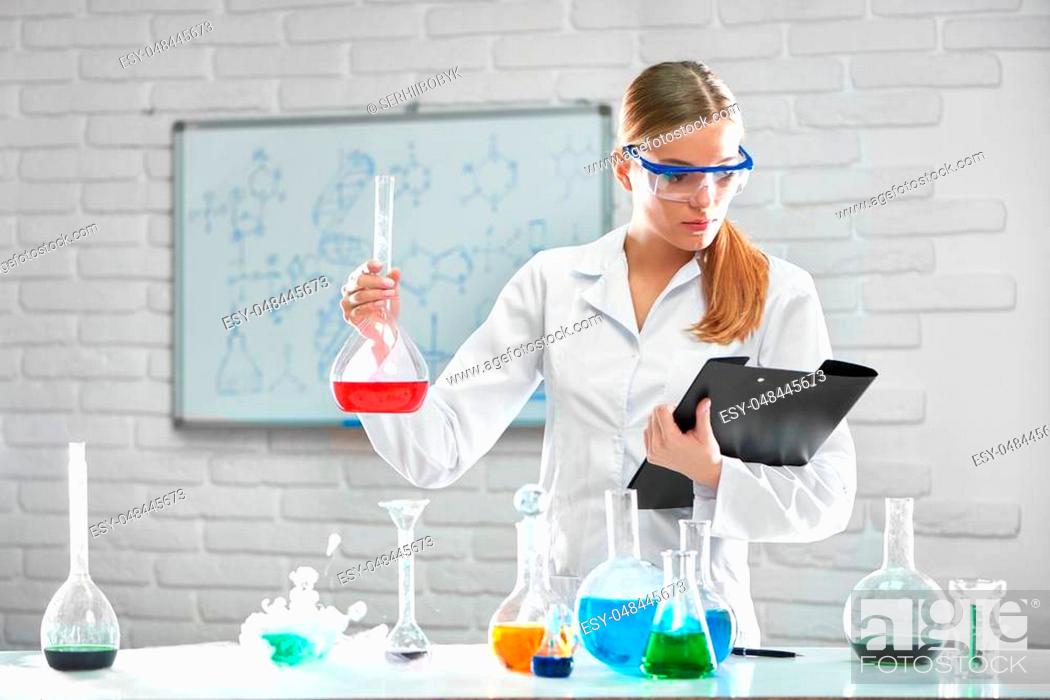 Stock Photo: Shot of a beautiful young female researcher in a labcoat and protective eyewear reading her notes on a clipboard holding a flask with red chemical liquid.