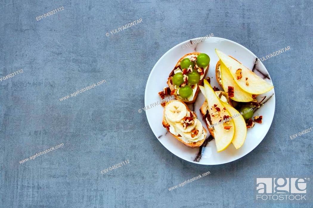 Stock Photo: Crostini with pear, cream-cheese, grapes, banana and nuts. Breakfast toasts or snack sandwiches on ceramic plate over grey stone background.