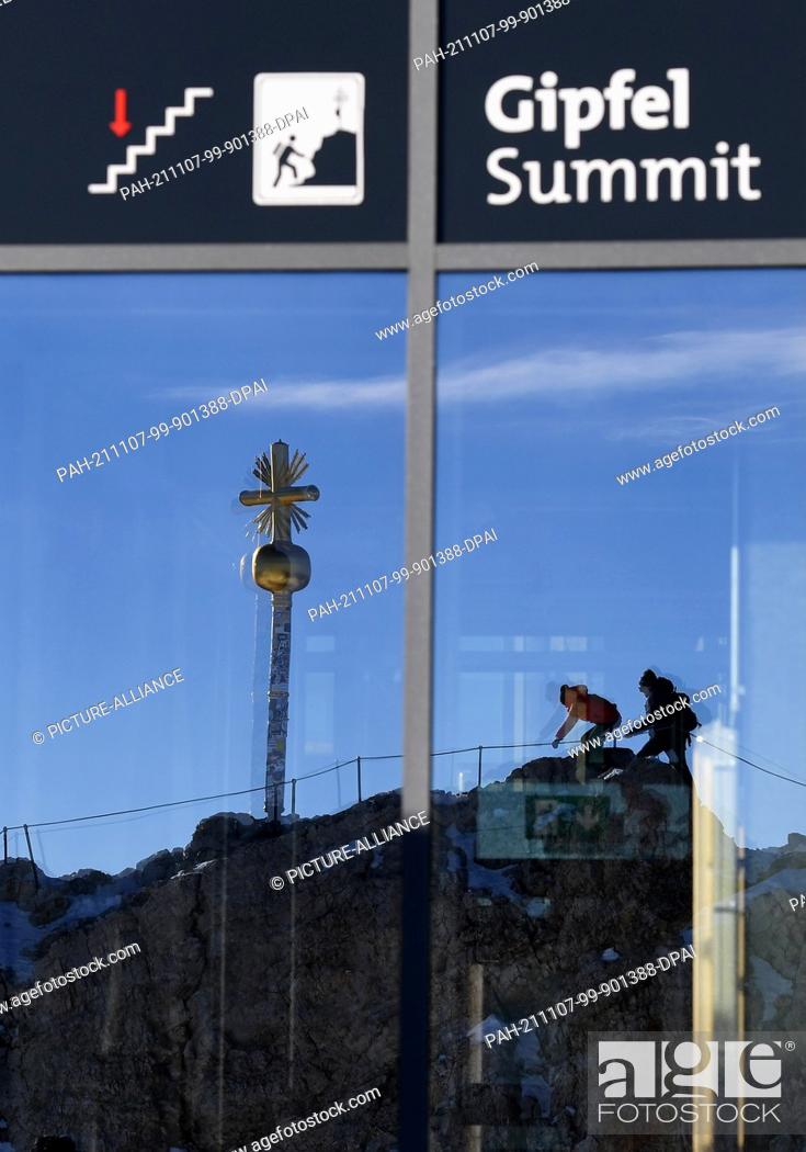 Stock Photo: 07 November 2021, Bavaria, Grainau: Two visitors climb the last few metres to the summit of the Zugspitze and are reflected in the mountain station of the.