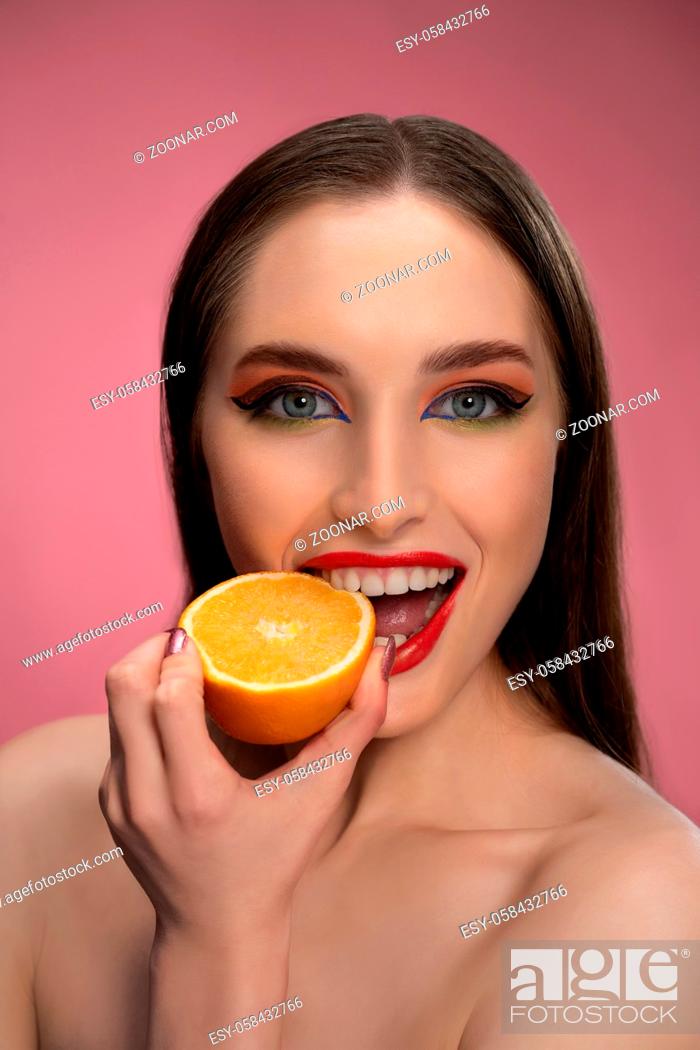 Stock Photo: Beautiful fashion girl holding orange sliced in half in her hand biting it looking at camera. Charming joyful funny lady with red lips and long hair isolated on.