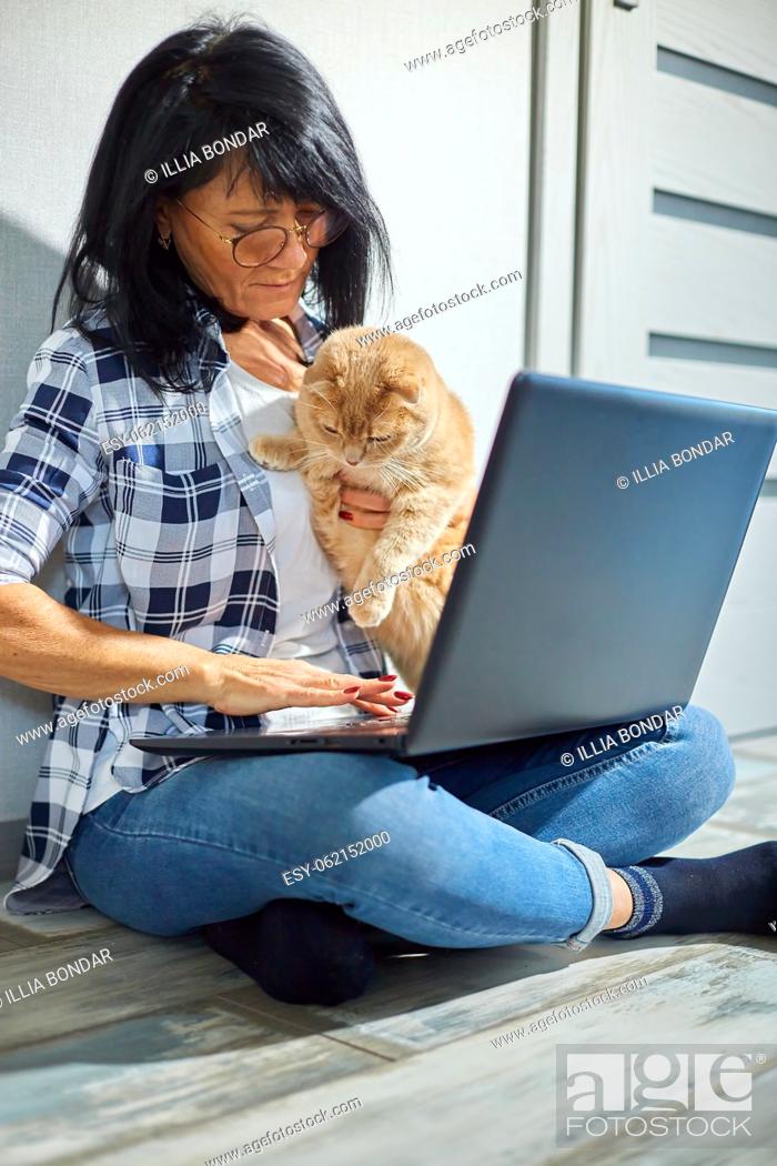 Stock Photo: Mature Woman searching vet website on laptop to register cat for veterinary consultation, Freelancer female typing on laptop sitting on the floor at home.