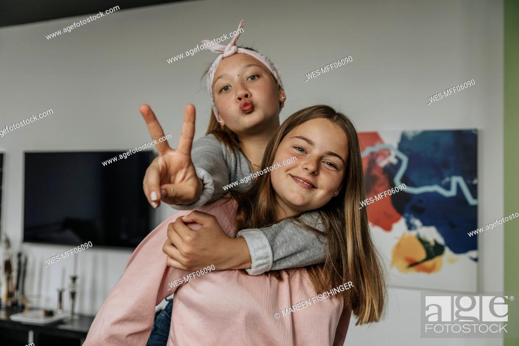 Stock Photo: Smiling girl piggybacking friend showing victory sign while standing at home.