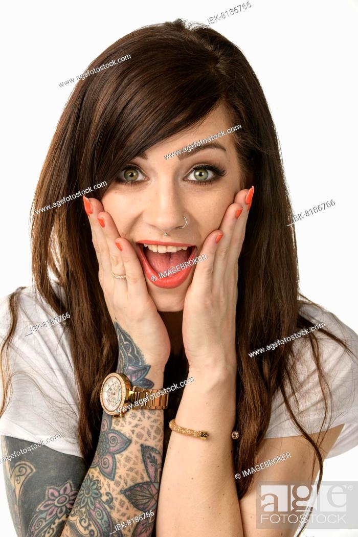 Stock Photo: Pretty young woman with long brunette hair, a white T-shirt and striking tattoos against a white background. With both hands she grasps her cheeks and shows an.